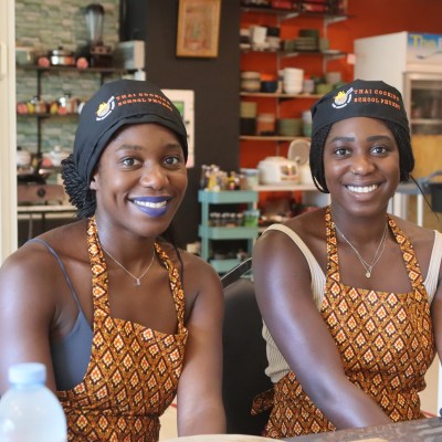 Two women in aprons and matching hats smiling at a table in a kitchen.