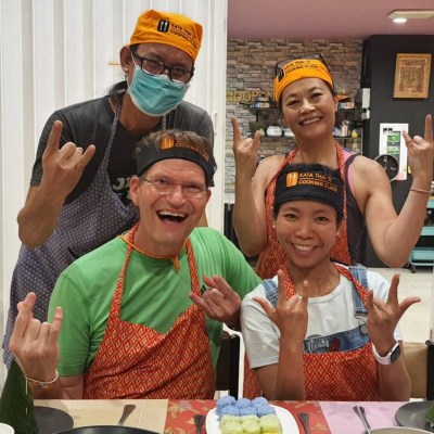 Four people wearing aprons smile and make hand gestures in a kitchen setting.